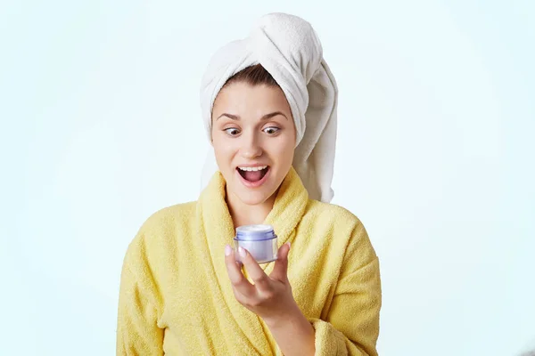 Portrait of young beautiful female being amazed by effect of new cosmetic cream, applies it on face after taking shower, has soft and delicate skin, wears domestic clothes, poses against white wall — Stock Photo, Image