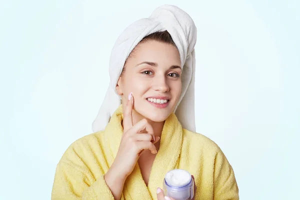 Mujer joven positiva con toalla en la cabeza, seca el cabello después de estar en el baño, aplica crema en la cara, demuestra su buen efecto, tiene una piel fresca, suave y saludable, aislada sobre fondo blanco —  Fotos de Stock