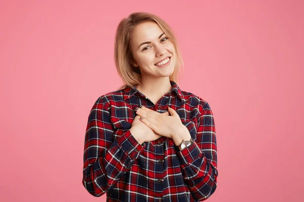 Romantico bella giovane femmina con piacevole sorriso splendente, tiene le mani sul petto, vestito con disinvoltura, dimostra la sua sensazione amichevole per le persone che la circondano, isolato su sfondo rosa — Foto Stock