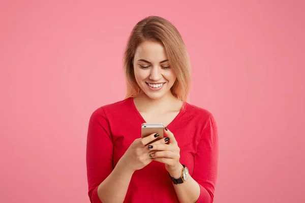 Waist up portrait of cheerful female in red sweater uses modern electronic device for online communication, surfes social networks, looks with glad positive expression into screen of cell phone — Stock Photo, Image