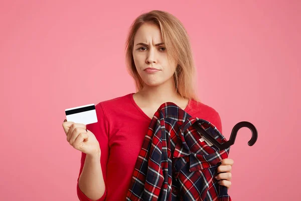 Photo of lovely woman with discontent expression goes shopping in fashionable boutique, chooses outfit, holds plastic card, has not enough money to pay for purchase, isolated on pink background — Stock Photo, Image