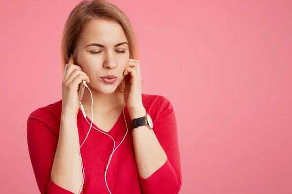 Wanita muda bergaya menggunakan ponsel dan earphone modern untuk mendengarkan musik atau lagu favoritnya dari playlist, menutup mata sebagai relaksasi perasaan dan kesenangan besar, terisolasi di atas latar belakang merah muda — Stok Foto