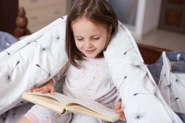 Horizontal shot of little girl reading under blanket with dandelion, having happy facial expression, looks exited, charming female child holding book in hands, reads fairy tale. Childhood concept. — Stock Photo, Image