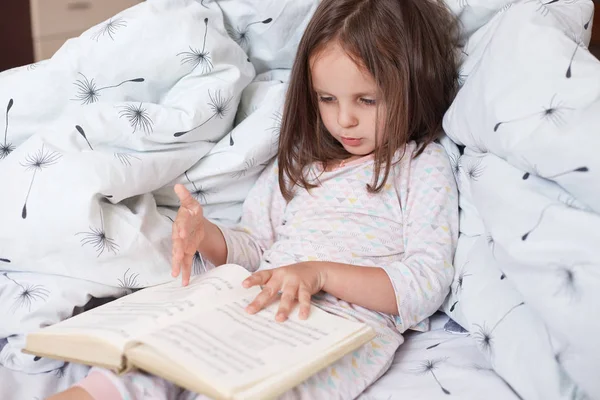 Horizontal shot of pretty little girl reading interesting book in her room while lying in bed under blanket with dandelion, looks concentrated and serious, cute dark haired female child rests at home. — Stock Photo, Image