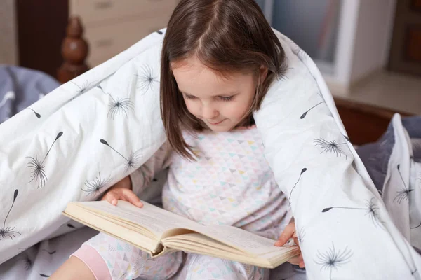 Portrait of charming child reading book at home, wrapped white blanket with dandelions, dark haired kid wearing pajama looking concentrated down on pages from her book. Children education concept. — Stock Photo, Image