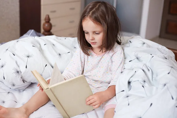 Horizontal shot of little girl sitting on her bed and reading interesting book, female child wearing white pajama, spending spare time in her cosy room, looks concentrated. Children education concept. — Stock Photo, Image