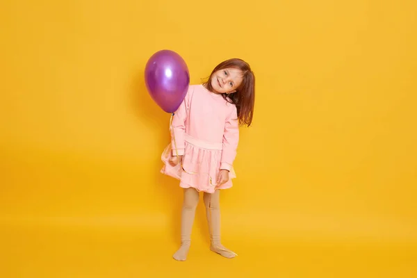 Tiro horizontal de menina bonito detém balão isolado sobre fundo amarelo, criança feminina usa vestido rosa, criança com cabelo escuro, olha sorrindo diretamente para a câmera, tem aniversário. Conceito de infância . — Fotografia de Stock