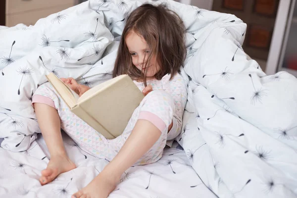 Indoor shot of child reading book in bed under blanket with dandelion. Female kid spending time in cozy bedroom. Little girl doing homework before sleepping, reading fairytale. Childhood concept. — Stock Photo, Image