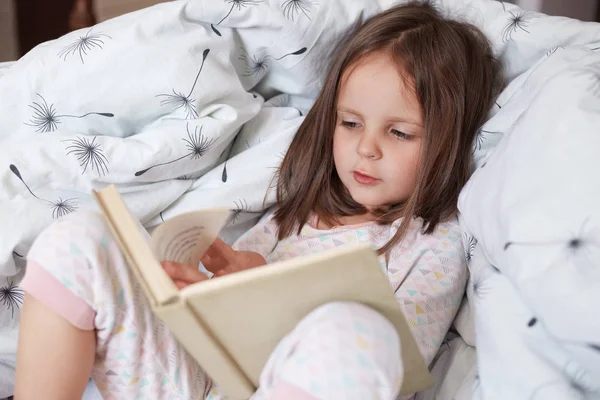 Hirizontal shot of happy girl lying in bed on blanket, reading interesting book,has concentrated facial expression, female child with dark hair likes to read fairy tales in morning. Childhood concept. — Stock Photo, Image