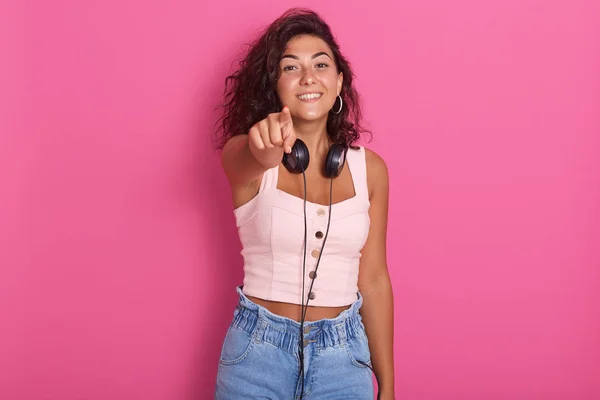 Närbild porträtt av söt flicka isolerad över rosa studio bakgrund, poiting med pekfingret till kamera, charmig kvinna bär rosentopp och jeans, hacing mörkt hår, flicka uttrycker lycka. — Stockfoto