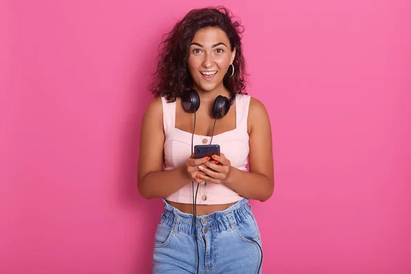 Retrato de una joven hermosa y feliz sosteniendo el teléfono en la mano y sonriendo, usando ropa casual, posando aislada sobre un fondo rosado, mujer con cabello ondulado oscuro, expresando asombro . — Foto de Stock