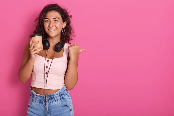 Studio shot of pleasant looking positive woman with satisfied facial expression, holding disposable cup of hot beverage, dressed casually, points aside, posing isolated over pink backround. Copy space — Stock Photo, Image