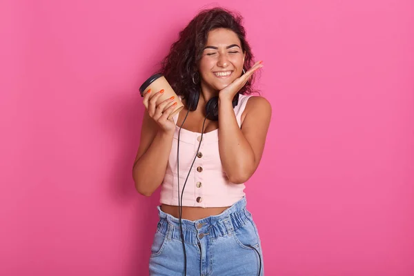 Imagen de la hermosa mujer caucásica joven y complacida, sosteniendo café caliente para llevar, usando ropa casual, de pie sobre fondo rosa, mujer disfrutando de un buen descanso y manteniendo la mano cerca de la mejilla . — Foto de Stock