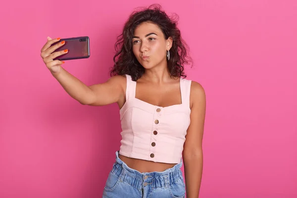 Горизонтальный снимок милой брюнетки в розовом топе и джинсах, делающей селфи на смартфоне на розовом фоне, показывающей жест поцелуя, женщины с темными волнистыми волосами. Технологическая концепция . — стоковое фото