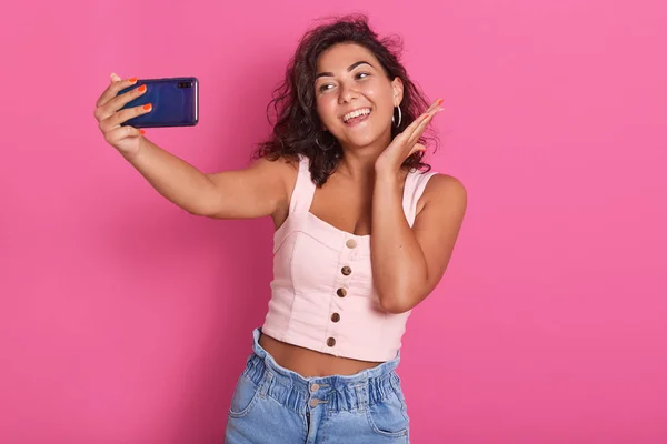 Tutup potret gadis muda yang lucu, dengan rambut hitam, mengenakan atasan mawar, membuat selfie pada ponsel pintarnya terisolasi di latar belakang merah muda, melihat layar perangkat dengan senyum bahagia. Konsep teknologi . — Stok Foto