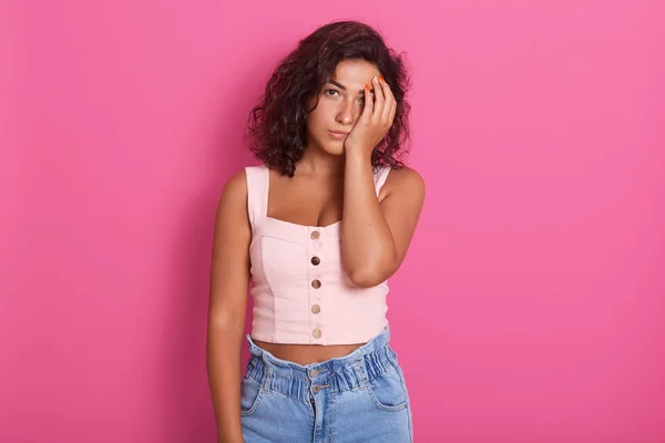 Horizontal shot of young winsome woman covers face with hand, has sleepy tired displeased facial expression, feels apathy, wearing stylish clothing, posing isolated over pink studio background.