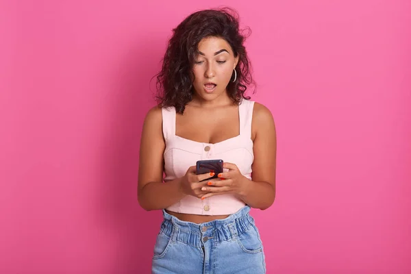 Portrait of shocked young woman holds smart phone, wearing stylish clothing, opened mouth with surprisment, looking her device screen, has astonished face expression, isolated on pink background. — Stock Photo, Image