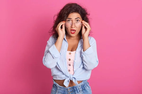 Fabulosa morena hembra con expresión aturdida, mira a través de las gafas, mantiene la boca abierta, ve algo sorprendente, posando aislada sobre fondo rosa, expresando asombro. Personas emociones . — Foto de Stock