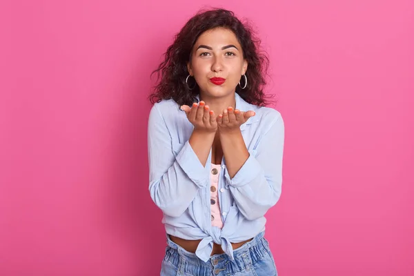 Gros plan portrait de jolie femme brune envoyant un baiser d'air à la caméra, mannequin posant isolé sur fond rose, femme séduisante portant une chemise bleue et un jean, ayant une manucure et un maquillage orange . — Photo