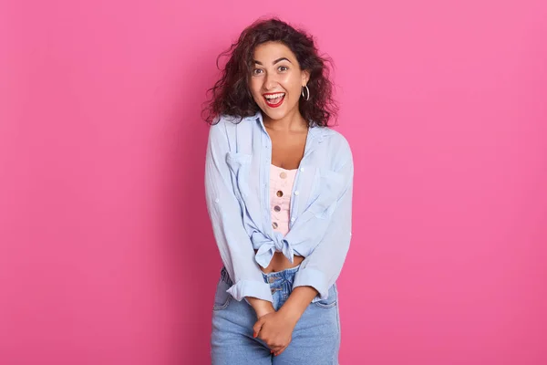 Imagen de una joven guapa hembra con cabello ondulado oscuro, vistiendo una camisa azul casual, maquillándose, mirando sonriendo directamente a la cámara, riendo, posando aislada sobre un fondo de srtudio rosa. Personas emociones —  Fotos de Stock