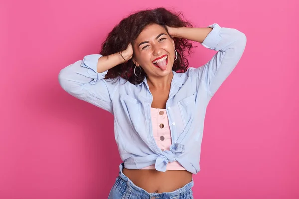 Retrato de chica encantadora con cabello ondulado oscuro, mostrando su lengua, mirando directamente a la cámara aislada sobre el fondo rosa, mujer con camisa azul, muestra el vientre desnudo, mantiene las manos en la cabeza, divirtiéndose . —  Fotos de Stock