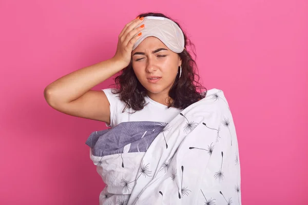 Horizontal shot of sleepy brunette young women with sleeping eye mask on forehead, wrapped in blanket, posing isoalted over pink background, keeps eyes closed and hand on head. Healthy care concept.