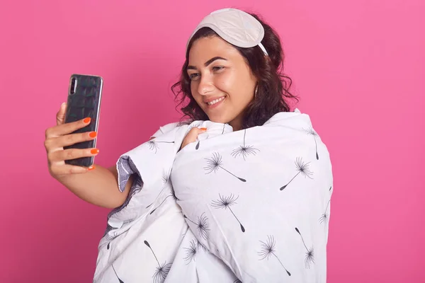 Indoor shot of lovely young woman sleep mask and wrappedwhite blanket, taking selfie during bedtime against pink background, adorable girl having time in home in morning before going for work . — Foto de Stock