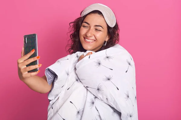 Portrait of smiling woman with blanket, adorable female with charming smile making selfie via her modern smartphone, looking at her device screen, making fun in morning, isolated over pink wall. — Stock Photo, Image