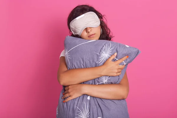 Front wiev of good looking young woman leans on gray pillow, wearing pyjama and eye mask, standing against pink studio background, does not want to wake up early in morning, resing at weekends. — Stock Photo, Image