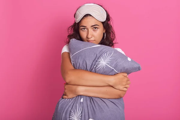 Studio shot of good looking young woman leans on pillow, winsome lady wearing pyjamas and eye mask, stands against pink background, has sleepy expression, posing early in morning. Rest concept. — ストック写真