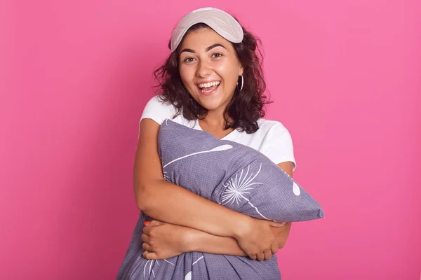 Relaxed sensual happy young woman hugging gray pillow, looking at camera with charming smile, standing isolated over pink background, expressing happyness, female wearing pajama and sleeping mask. — 스톡 사진