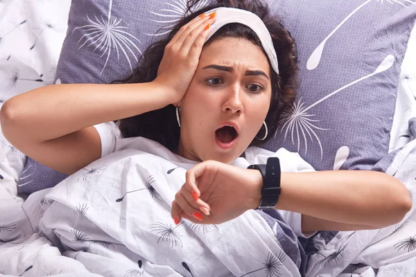 Portrait of young woman lying in bed on pillow and under blanket with dandelion, being at home in her cosy room, keeps mouth opened, looking at watch with astonish facial expression, being late. — Stock fotografie