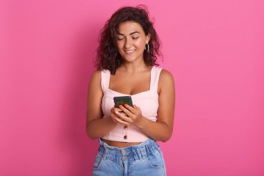 Elinde cep telefonu olan neşeli bir kadın, elinde akıllı bir telefon tutan ve gülümseyen bir kadın, pembe bluz ve kot pantolon giyen bir kadın, sosyal ağları kontrol ediyor..