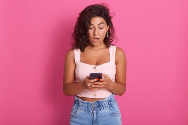 Şok olmuş genç bir kadının portresi şık giysiler içinde akıllı bir telefon tutuyor. Ağzını şaşkınlıkla açıyor. Cihaz ekranına bakıyor..