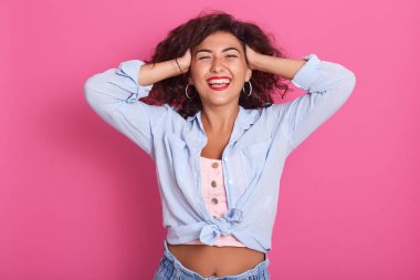 Mavi gömlekli ve kot pantolonlu güzel bir kadının portresi, çıplak göbeğini gösteriyor, tok bir gülümsemeyle poz veriyor, pembe arka planda tek başına duruyor, mutluluğunu ifade ediyor, elini başının üstünde tutuyor..