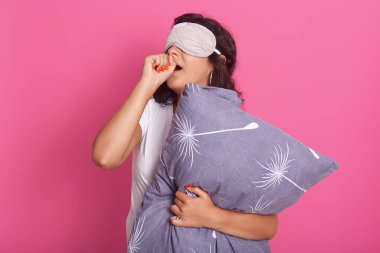 Indoor shot of attractive young woman standing against pink studio wall, posing in sleeping mask on her eyes and pajamas, holding pillow, covering mouths with hands and yawning. Morning time concept.