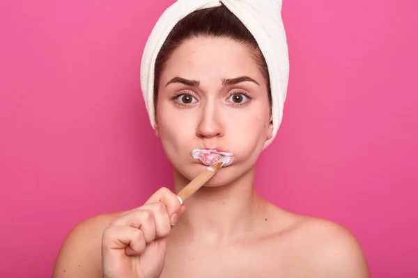 Retrato de una atractiva mujer caucásica con cabello oscuro y toalla blanca en la cabeza, posando aislada sobre la pared del estudio rosa, mirando a la cámara y cepillándose los dientes. Concepto procedimiento matutino . — Foto de Stock