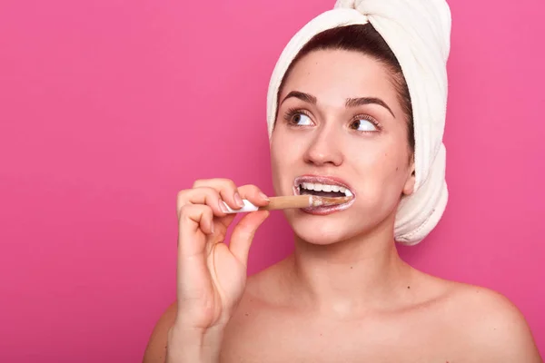 Retrato de cerca de una joven adorable de pie aislada sobre el fondo del estudio rosa y cepillándose los dientes, usando una toalla blanca, teniendo una expresión facial reflexiva, sueña con algo . — Foto de Stock
