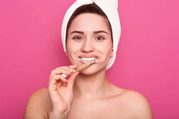 Afbeelding van mooi jong meisje verpakt witte handdoek, het reinigen van haar tanden, ziet er gelukkig uit, in goed humeur, het doen van ochtendprocedures in de badkamer, poseren met ontbloot schouders tegen roze studio muur. — Stockfoto