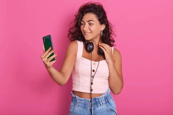 Wanita perhatian bersenang-senang dan membuat foto selfie menggunakan telepon, wanita remaja mengenakan headphone hitam di sekitar leher dan atas kemerahan, gadis mendengarkan musik di dalam ruangan, terisolasi di atas latar belakang merah muda . — Stok Foto