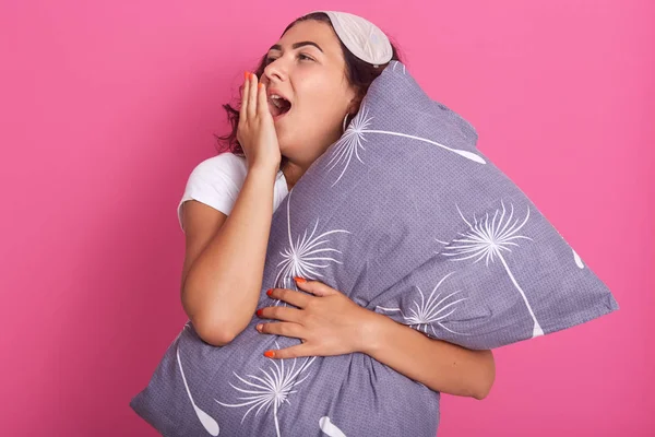 Portrait of attractive woman yawning as has sleepy expression, wearing domestic clothes, carries soft gray pillow, poses against rosy background. People, rest, comfort, tiredness, sleeping concept. — Φωτογραφία Αρχείου