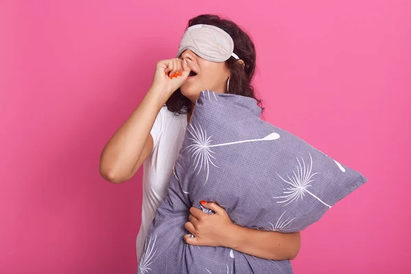 Indoor shot of attractive young woman standing against pink studio wall, posing in sleeping mask on her eyes and pajamas, holding pillow, covering mouths with hands and yawning. Morning time concept. — Φωτογραφία Αρχείου