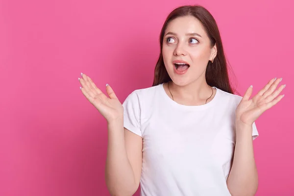 Menina incrível vestindo camisa branca casual t mantendo a boca bem aberta e espalhando as mãos, olhando para o lado, modelo posando isolado sobre fundo de parede rosa no estúdio. Conceito de emoções sinceras . — Fotografia de Stock