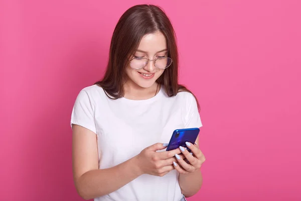 Horizontal shot of adorable teen with long hair, holding modern cell phone, scrolls through social networks, has cheerful expression, wears spectacles and casual t shirt, isolated over pink background — Stock Photo, Image