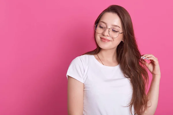 Portret szczęśliwej młodej, ładnej, brązowłosej dziewczyny sytands uśmiechającej się i patrzącej na bok, dotyka włosów, pozując na różowym tle, wygląda tysięcznie, śni o czymś. Copyspace na reklamę — Zdjęcie stockowe