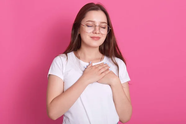 Close up portrait of thankful young woman wearing white casual t shirt posing isolated over pink studio background, standing with closed eyes, keeps hands at heart chest, feels gratitude, grateful.