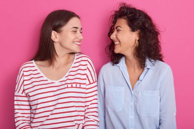 Pembe arka planda poz veren iki güzel genç kadının yakın portresi, günlük kıyafetler giyen, ayakta duran ve birbirlerine gülümseyen koyu renk saçlı kadınlar. Arkadaşlık kavramı.