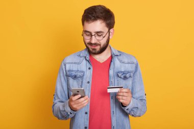 Studio shot of young european bearded man holding credit card and smartphone, standing with calm facial expression over yellow background, spending money from his account. Online shopping concept.
