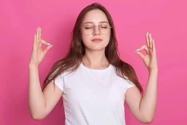 Concepto de relajación y meditación. Hermosos vestidos de mujer blanca camiseta casual meditando con los ojos cerrados, relajando el cuerpo y despejando la mente, preparándose para un nuevo día feliz, aislada sobre rosa . — Foto de Stock