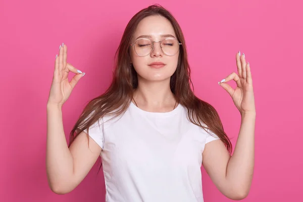 Avkoppling och meditation koncept. Vackra kvinnliga klänningar vit casual t skjorta meditera med ögonen stängda, avkopplande kropp och clearing sinne, förbereda sig för ny lycklig dag, isolerade över rosa. — Stockfoto